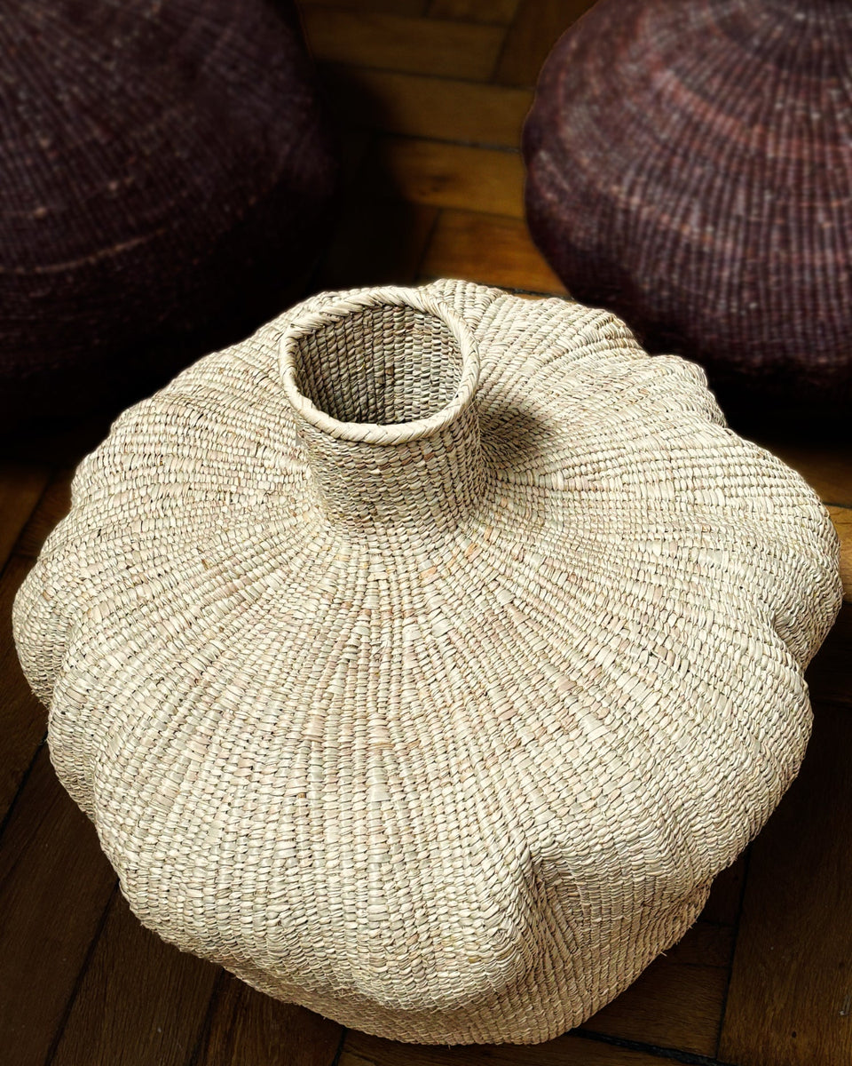 A hand-woven natural fiber vase with a flared, rounded body and a short neck, resting on a wooden floor with other woven baskets in the background.