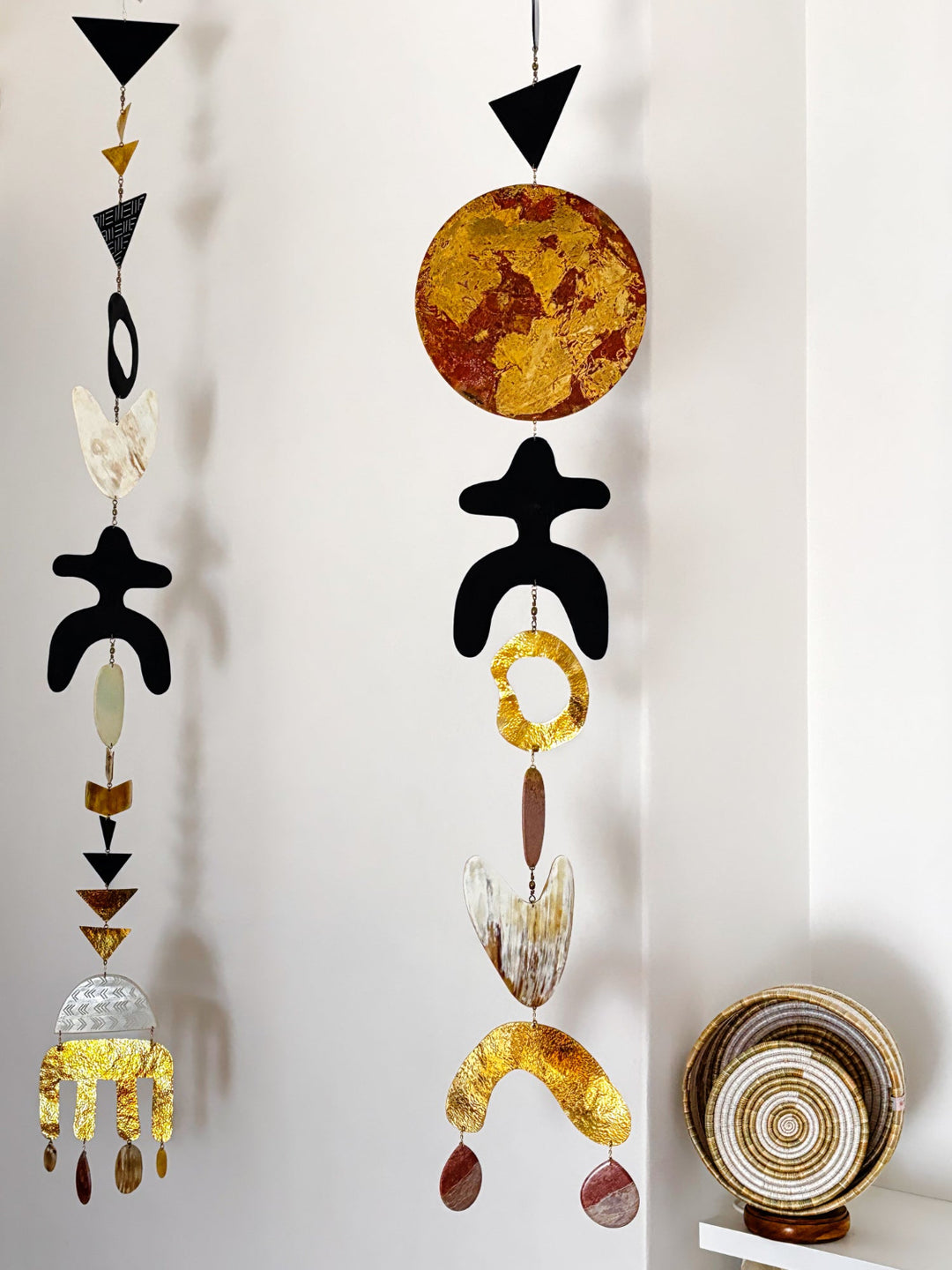 Two abstract metal wall hangings with black, gold, brown, and white organic shapes suspended against a white wall, with a decorative bowl on a shelf to the right.