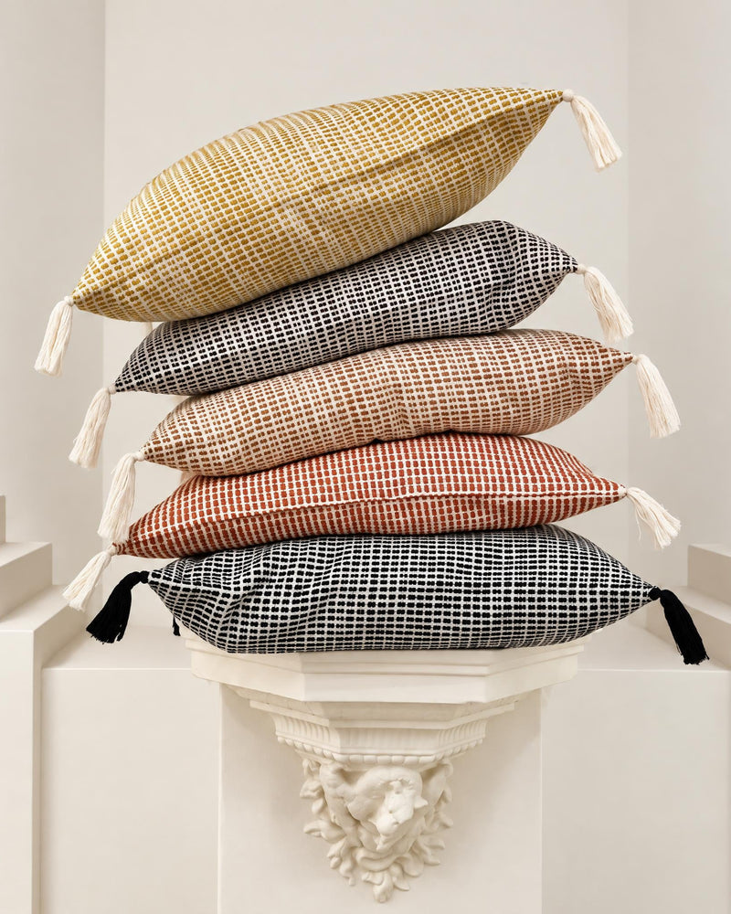 Five woven throw pillows in different colors and patterns are stacked on a white decorative pedestal in a bright, minimalist room. The pillows feature small tassels on the sides.
