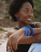 A woman outdoors wearing bright blue hoop earrings and matching thick blue bangles on her wrist, with a blurred natural background.