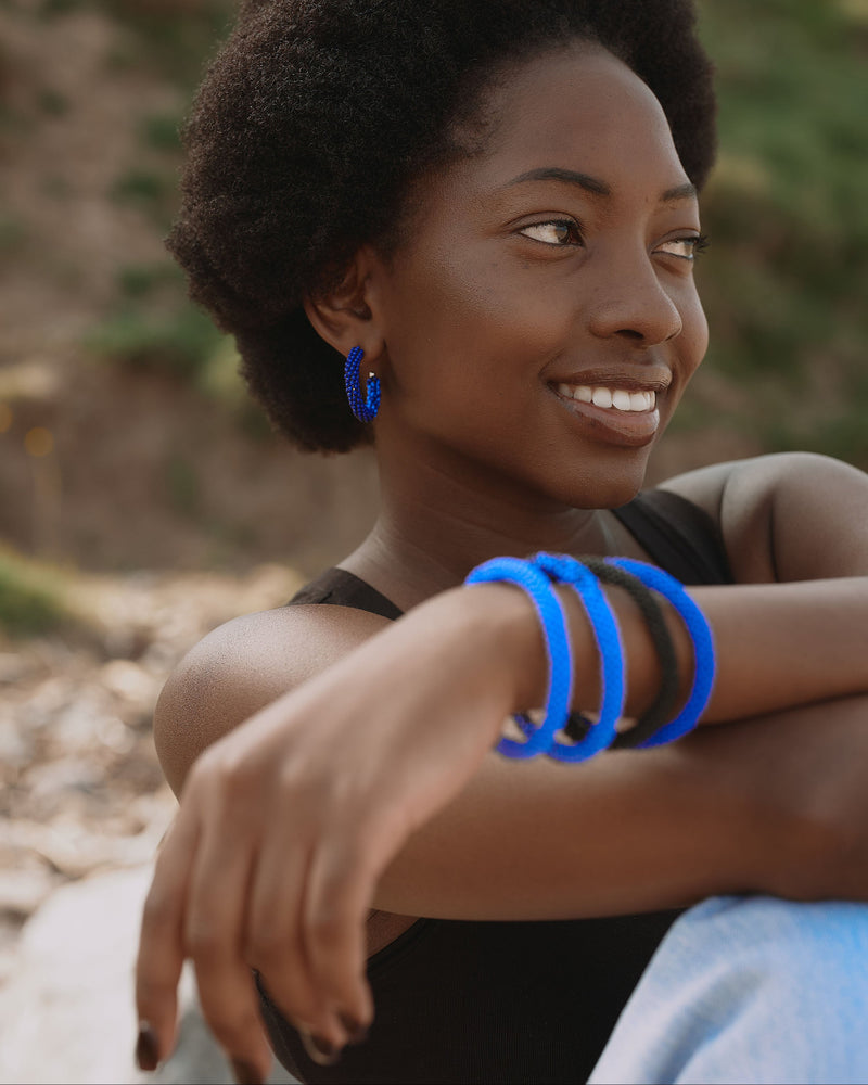 A woman outdoors wearing bright blue hoop earrings and matching thick blue bangles on her wrist, with a blurred natural background.