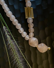 A beaded necklace made of pale peach-pink translucent glass beads, featuring a textured gold rectangular accent and a large irregular bead at the end, styled with dried grass against a dark textured background.