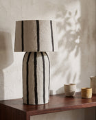 A table lamp made from paper pulp with a drum shade and a matching cylindrical base, featuring vertical black stripes, displayed on a dark wood console table against a textured off-white wall.