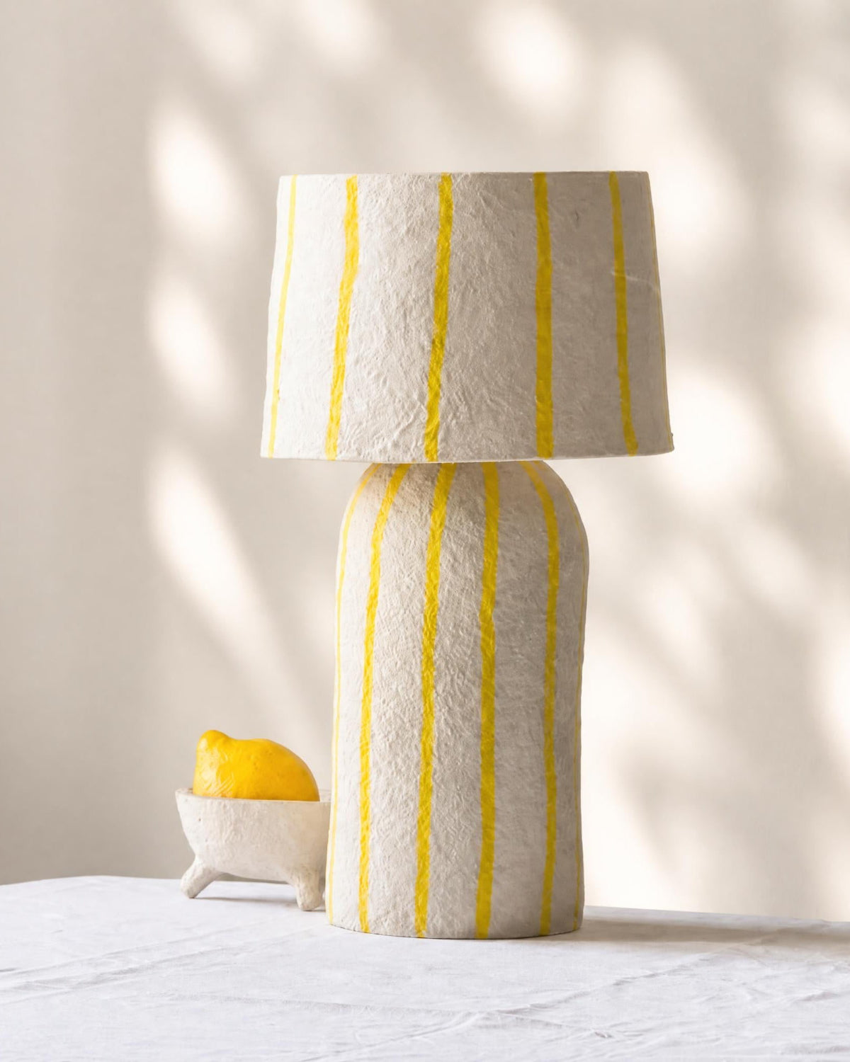 A table lamp made from paper pulp with an off-white body and matching drum shade, decorated with vertical yellow stripes, set on a white tablecloth next to a small bowl of lemon under soft leaf shadows.